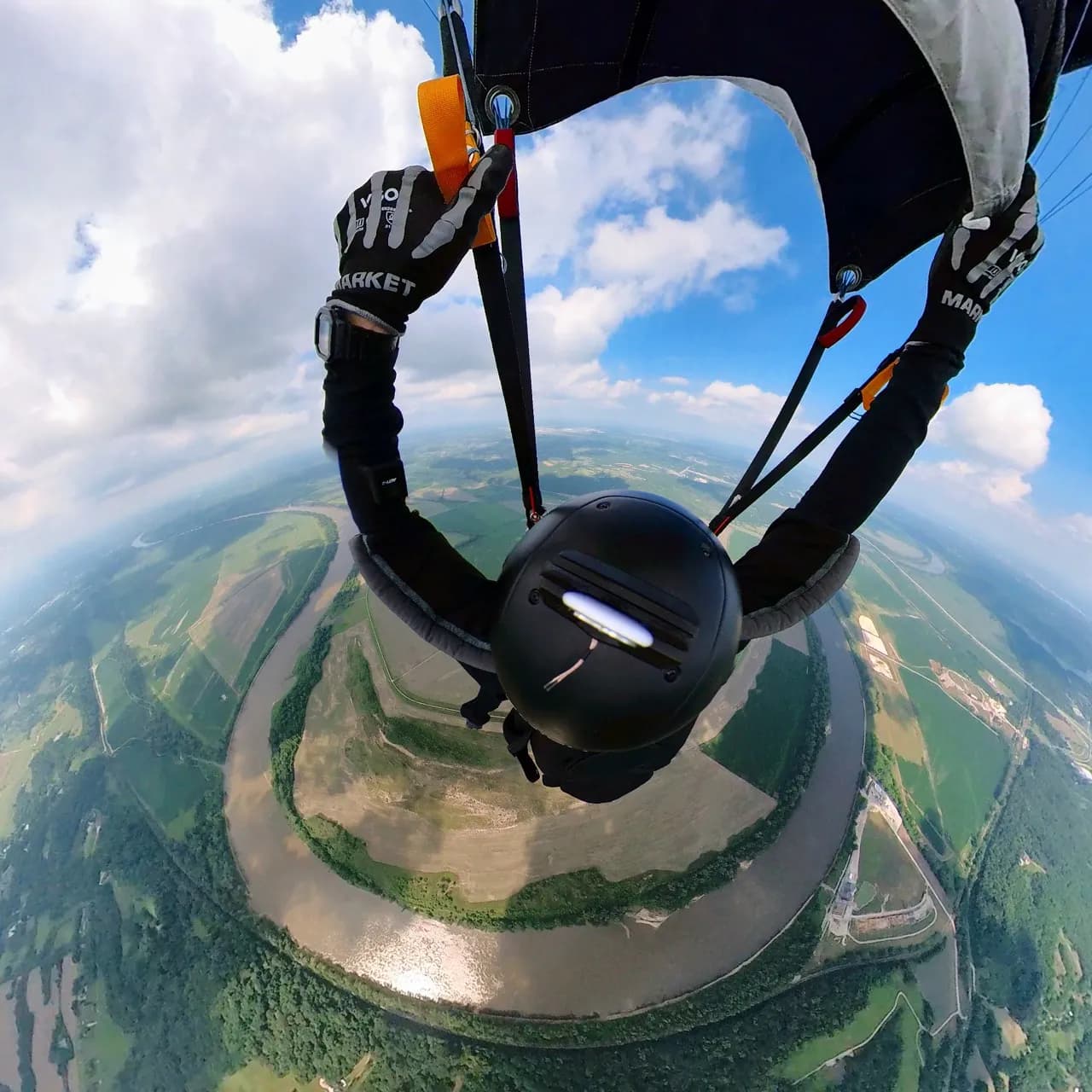 Wide-angle view from under an open parachute, looking down past the harness at a winding river and farmland far below.